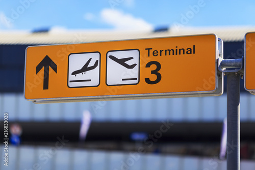 Yellow sign board with arrow, arrival and departure plane icons and 'Terminal three' text on blurred airport building background