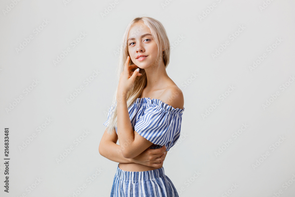 Naklejka premium Calm and friendly beautiful blonde girl in summer outfit is smiling posing at camera, over white background. Emotions, gestures and facial expression