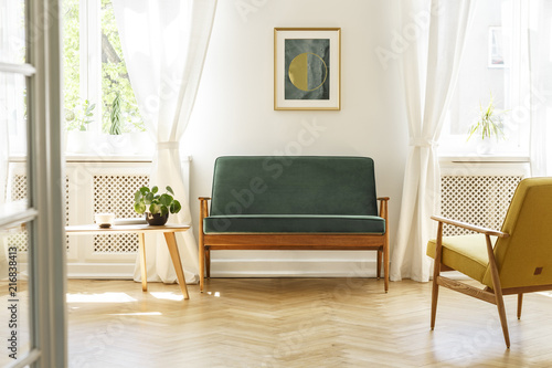 Green sofa with dark, wooden frame and a comfy yellow armchair in a white retro living room interior with natural light coming through big windows. Real photo.