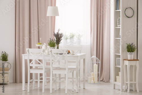 Lamp above table and white chairs in pink dining room interior with plants and drapes. Real photo © Photographee.eu
