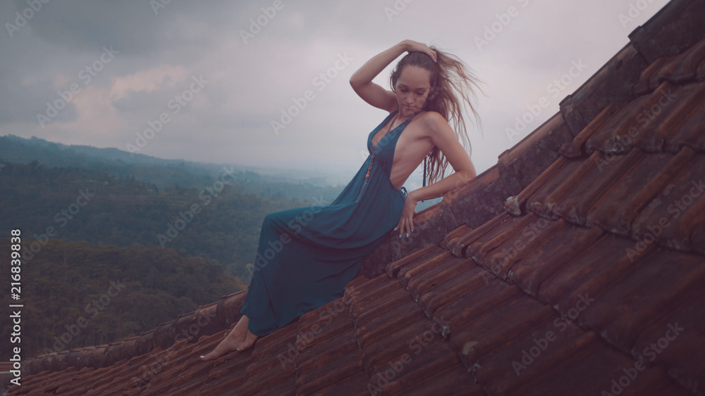 Aerial drone view of beautiful woman in long blue dress sitting on tiled red roof of the house against amazing mountain view and cloudy sky background - obrazy, fototapety, plakaty 