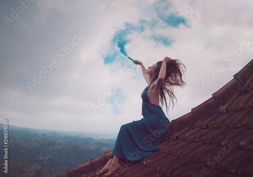 Beautiful woman in long dress sitting with blue colored smoke on tiled red roof of the house against amazing mountain view and cloudy sky background