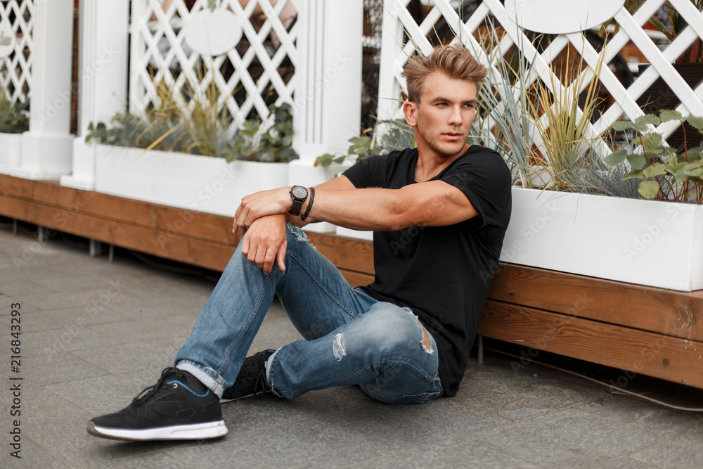 stylish young man with a hairstyle in a fashionable black T-shirt with blue jeans in sneakers sits on the beach near a white wooden fence - obrazy, fototapety, plakaty 