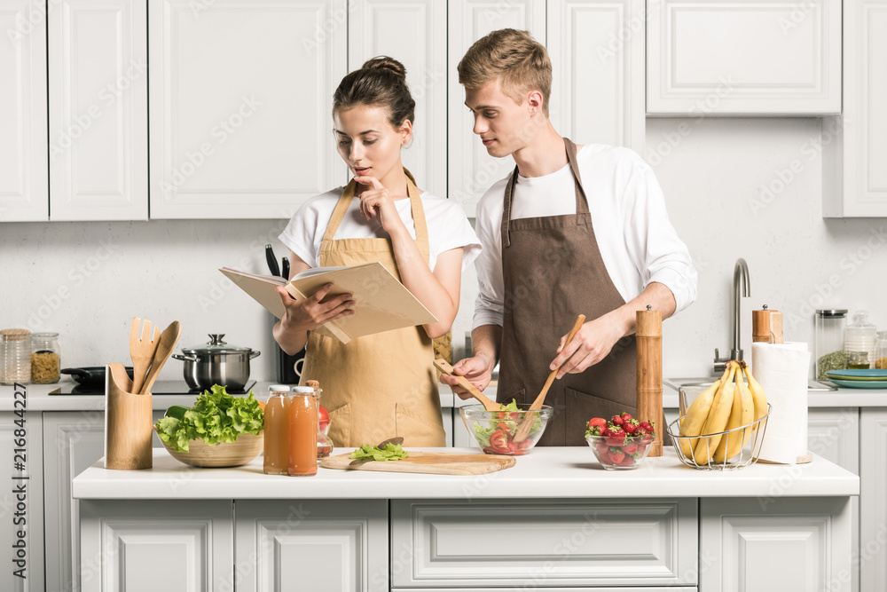 young couple cooking salad and reading recipe in cookbook in kitchen young couple cooking salad and reading recipe in cookbook in kitchen