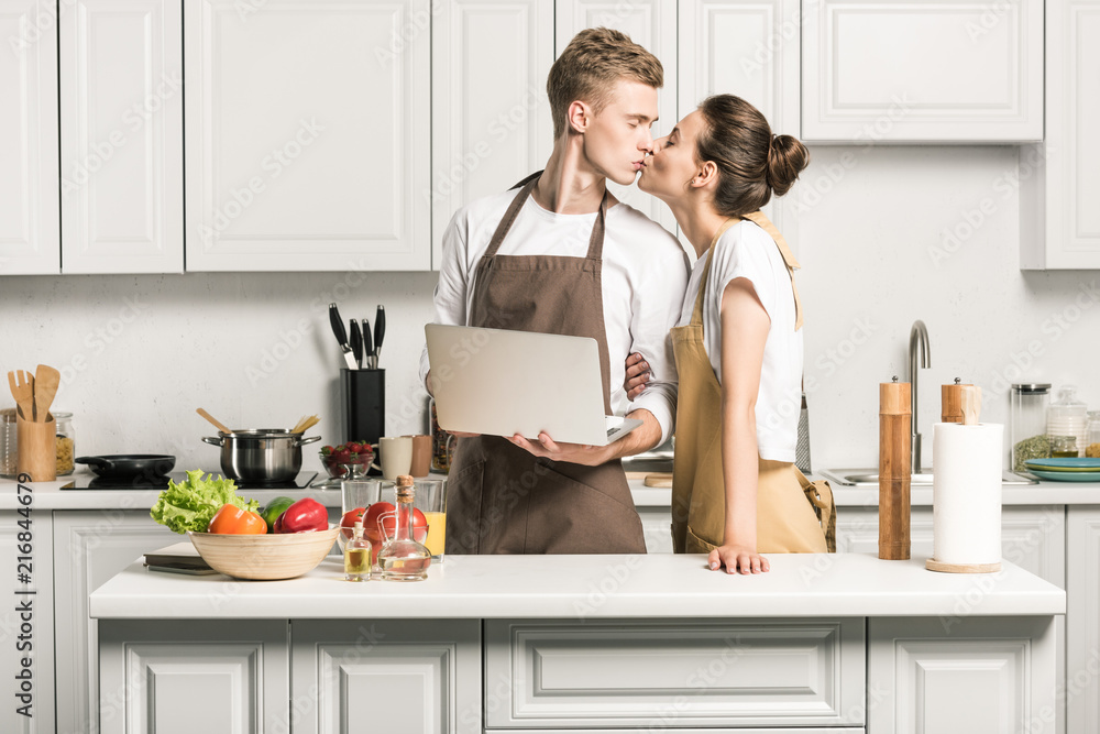 young couple kissing in kitchen and holding laptop