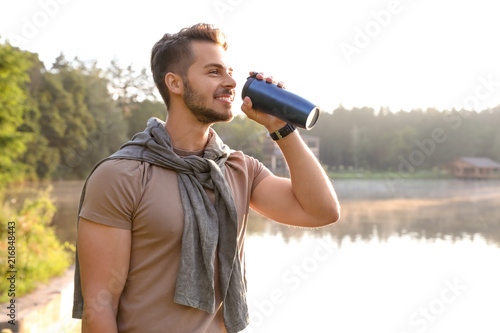 Young man drinking water on shore of beautiful lake. Camping season