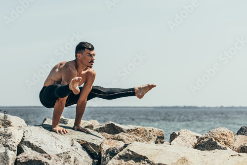 sporty shirtless man doing arm balance on rocky seashore