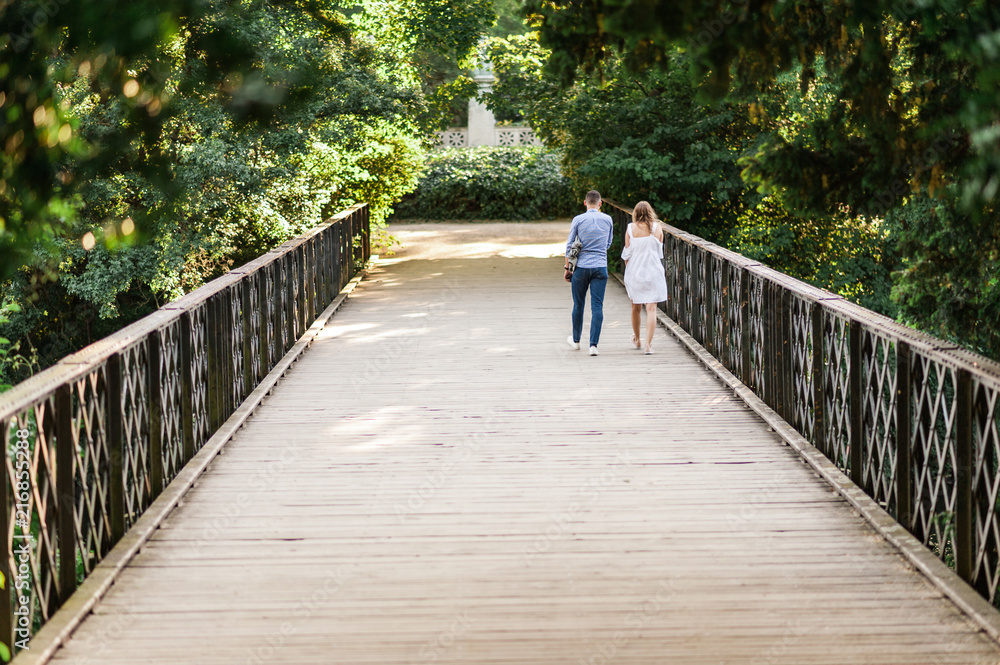Young couple walking along peaceful park scenery on a beautiful summer day. Green canopy of trees and a metal bridge above the water of a pond. Concept of hygge. Young couple walking along peaceful park scenery on a beautiful summer day. Green canopy of trees and a metal bridge above the water of a pond. Concept of hygge.