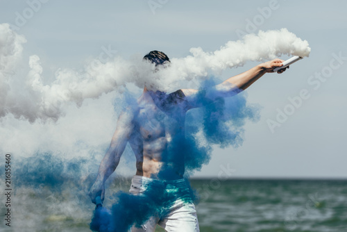shirtless man with blue and white smoke sticks in front of ocean