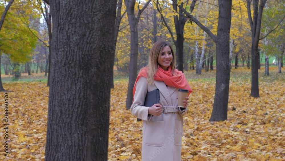 Woman Walks On The Autumn Park Holding A Folder Or A Laptop And Drinking Coffee Woman Walks On The Autumn Park Holding A Folder Or A Laptop And Drinking Coffee