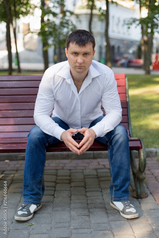 the guy is sitting on a bench in the Park. a man in a white shirt and jeans. young man. a man in a white shirt portrait of a man. business portrait. male portrai the guy is sitting on a bench in the Park. a man in a white shirt and jeans. young man. a man in a white shirt portrait of a man. business portrait. male portrai