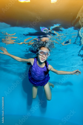 Funny little girl is swimming underwater in the pool on yellow light background in swimming glasses and purple dress. Portrait. Underwater photography. Vertical view