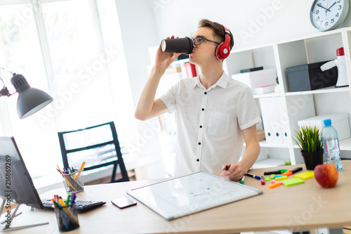 A young man with glasses and headphones drinks coffee. On the table in front of him lie a magnetic board and a marker.