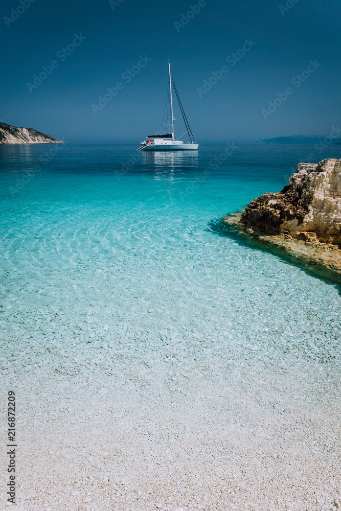 Beautiful azure blue lagoon with sailing catamaran yacht boat at anchor. Pure white pebble beach, some rocks in the sea