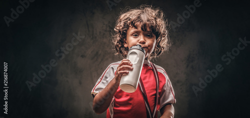 Little champion boy in sportswear with a gold medal drinking water from a bottle. Isolated on a dark textured background.