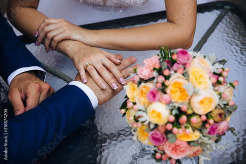 Close-up of a cropped frame of the bride and groom on their wedding day holding hands. The girl wears an engagement ring with a diamond, a French manicure of a bride, a wedding bouquet.