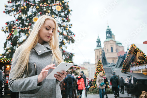 Photography A beautiful young blonde woman or tourist uses a tablet next to a Christmas tree during the Christmas holidays at the Old Town Square in Prague, Czech Republic
