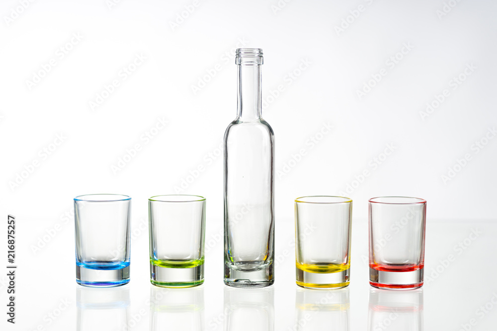 Four multicolored empty shot glasses and small bottle placed symmetrically on a white background. Four multicolored empty shot glasses and small bottle placed symmetrically on a white background.