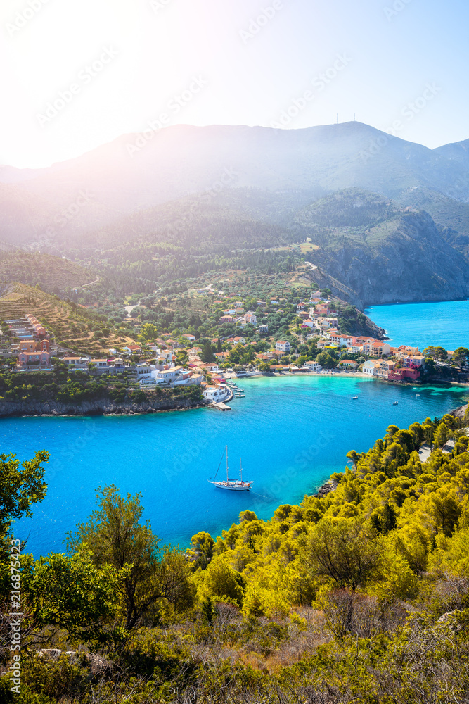 Assos village in morning light, Kefalonia. Greece. White lonely yacht in beautiful turquoise colored bay lagoon water surrounded by pine and cypress trees along the coastline