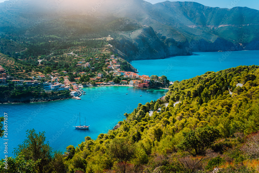 Panoramic view to Assos village Kefalonia. Greece. White lonely yacht in beautiful turquoise colored bay lagoon water surrounded by pine and cypress trees along the coastline