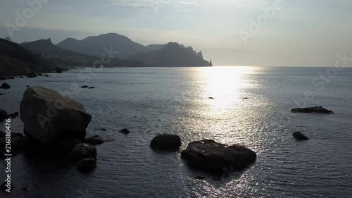 Flying backward over the sea surface with rocks and stones at sunrise. Mountains, rocks, stones in the wwater and sea waves. Aerial view of Kara Dag Mountain in Crimea. photo