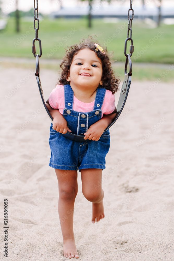 Portrait of happy smiling little latin hispanic toddler girl swinging on swings at playground outside on summer day. Happy childhood lifestyle concept. Toned with film pastel faded filters colors.