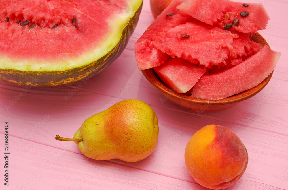 Pieces of watermelon in a plate and a watermelon, a pear, a peach on a pink wooden table Pieces of watermelon in a plate and a watermelon, a pear, a peach on a pink wooden table