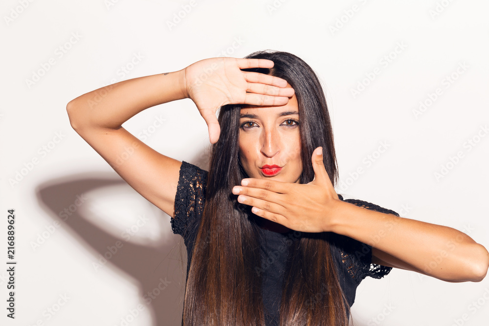 Fototapeta premium Young adult woman framing her face with her hands, making funny faces. In studio over a white background.