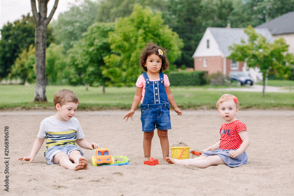 Portrait of three cute Caucasian and hispanic latin toddlers babies children sitting in sandbox playing with plastic colorful toys. Little girls and boy friends having fun together on playground. Portrait of three cute Caucasian and hispanic latin toddlers babies children sitting in sandbox playing with plastic colorful toys. Little girls and boy friends having fun together on playground.