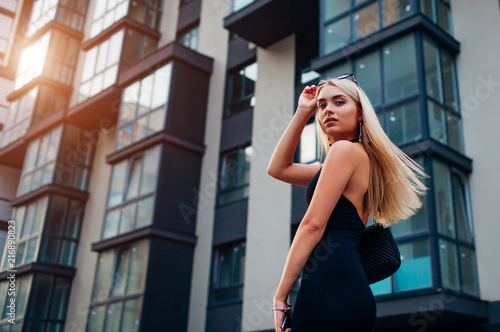Portrait of young blonde woman wearing stylish clothing and accessories by modern business center in city.