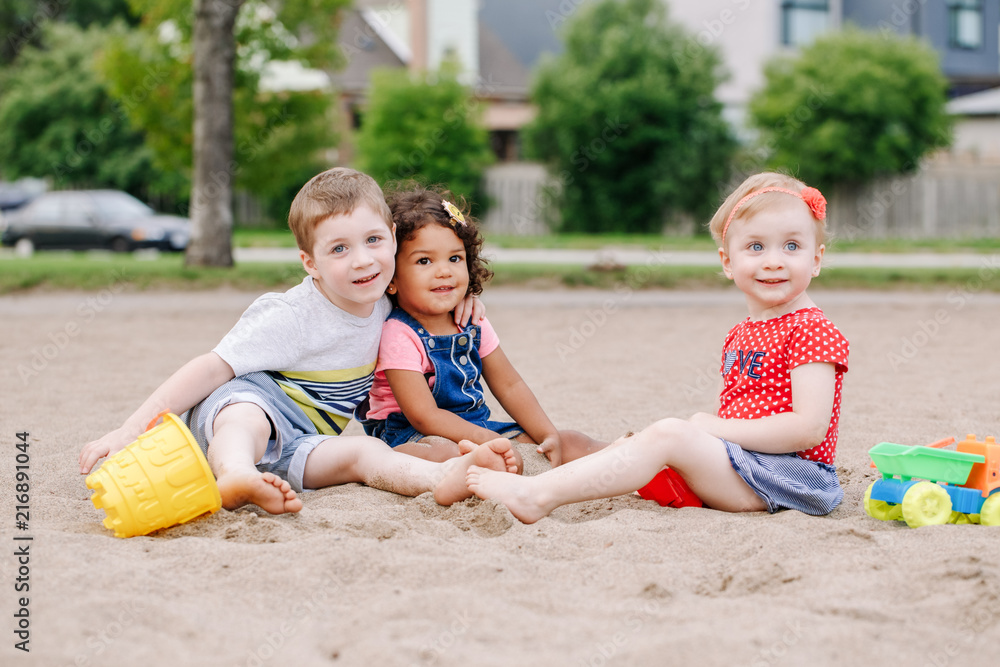 Portrait of three cute Caucasian and hispanic latin toddlers babies children sitting in sandbox playing with plastic colorful toys. Little girls and boy friends having fun together on playground. Portrait of three cute Caucasian and hispanic latin toddlers babies children sitting in sandbox playing with plastic colorful toys. Little girls and boy friends having fun together on playground.