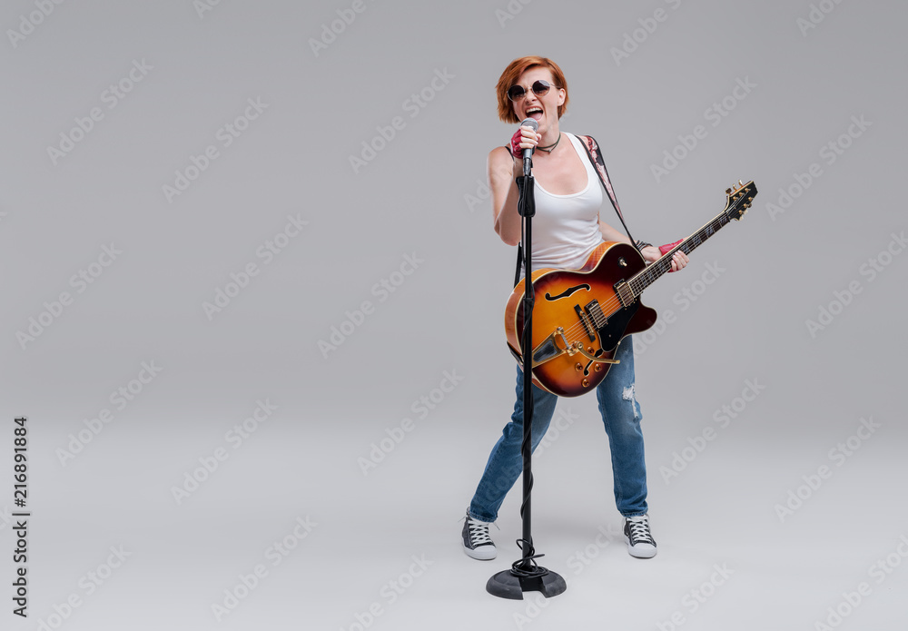 Young woman musician with an acoustic guitar in hand on a gray background. He laughs and plays rock and roll loudly. Full-length portrait. On the left there is space for text Young woman musician with an acoustic guitar in hand on a gray background. He laughs and plays rock and roll loudly. Full-length portrait. On the left there is space for text