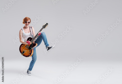 Young woman musician with an acoustic guitar in hand on a gray background. He laughs and plays rock and roll loudly. Full-length portrait. On the right there is space for text photo
