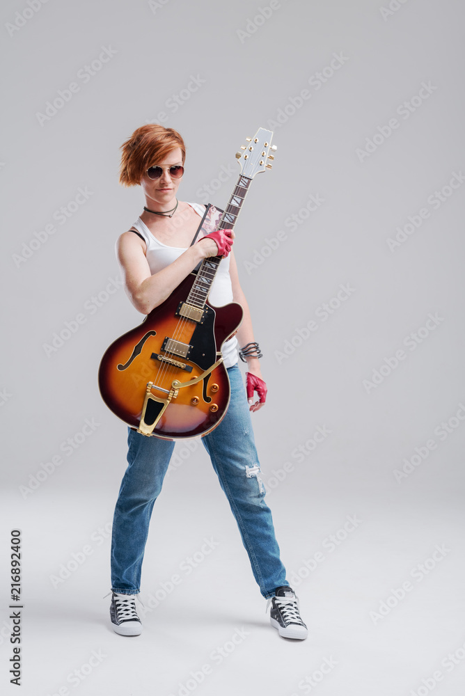 Fototapeta premium Young woman musician with an acoustic guitar in hand on a gray background. He laughs and plays rock and roll loudly. Full-length portrait