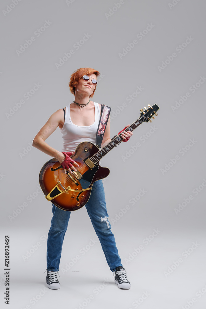 Young woman musician with an acoustic guitar in hand on a gray background. He laughs and plays rock and roll loudly. Full-length portrait