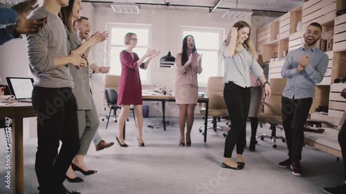 Happy European girl doing moonwalk dance at fun office party. Multiethnic business people celebrate success slow motion. photo