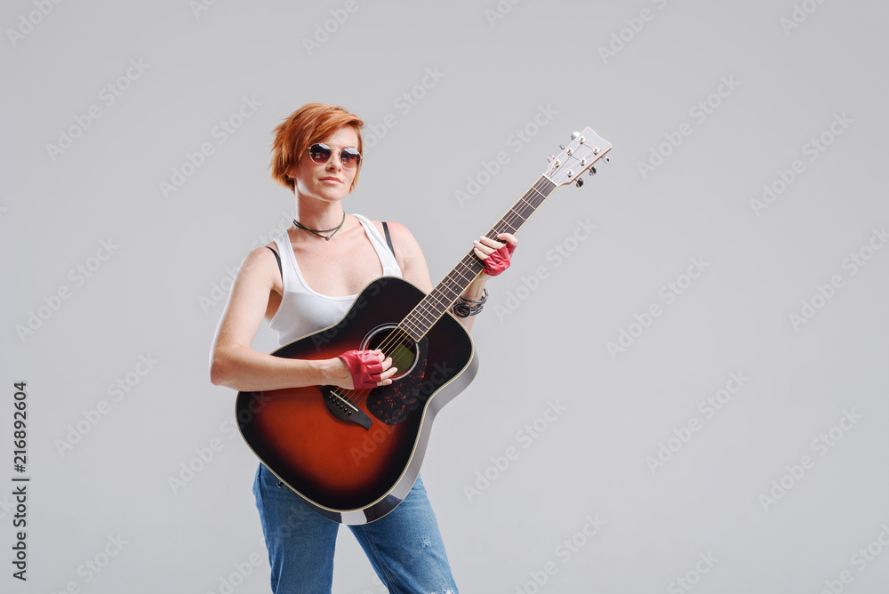 Young woman musician with an acoustic guitar in hand on a gray background. He laughs and plays rock and roll loudly. Young woman musician with an acoustic guitar in hand on a gray background. He laughs and plays rock and roll loudly.