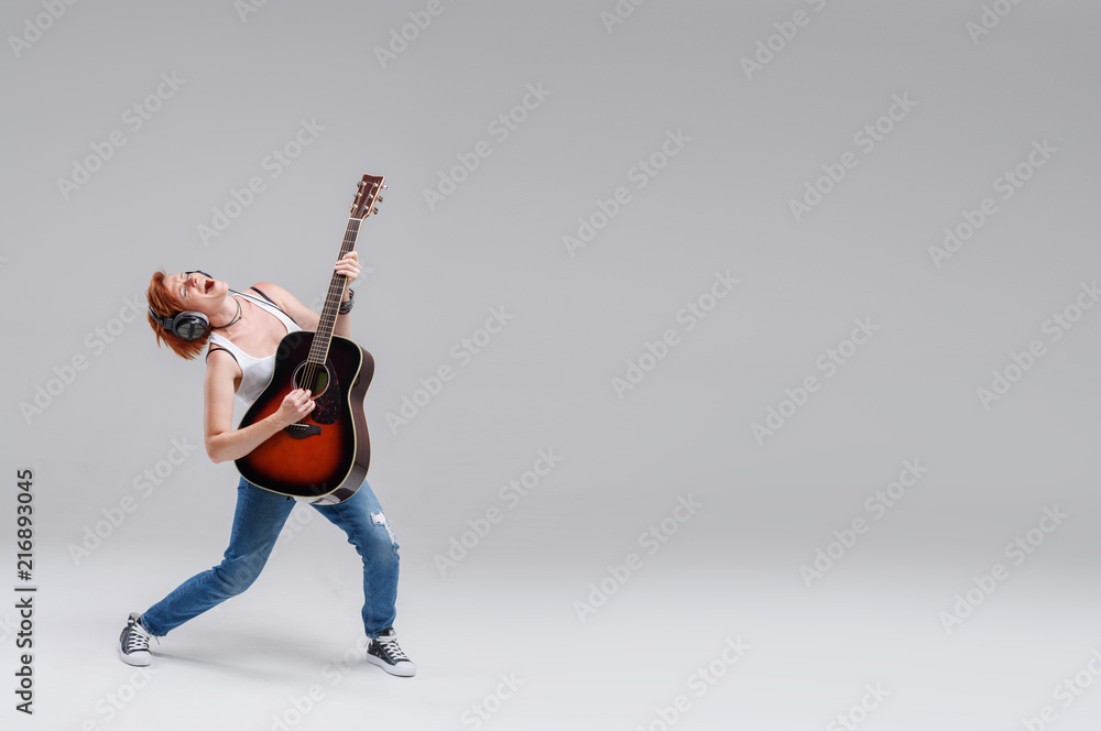 Young woman musician with an acoustic   guitar in hand and in headphones on a gray background. He laughs and plays rock and roll loudly. Full-length portrait. On the right there is space for text