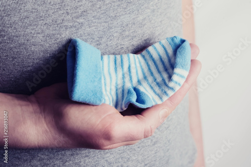 Small blue sock for the unborn baby in the belly of pregnant woman. Hands of happy woman holding little sock of child over gray t-shirt