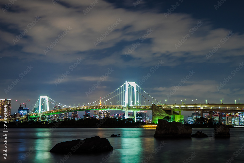 Obraz premium Rainbow bridge at night shot from Odaiba island, Tokyo, Japan. Dark rocks in the front stick out from the water, and scyscrapers illuminated with lights are in the background