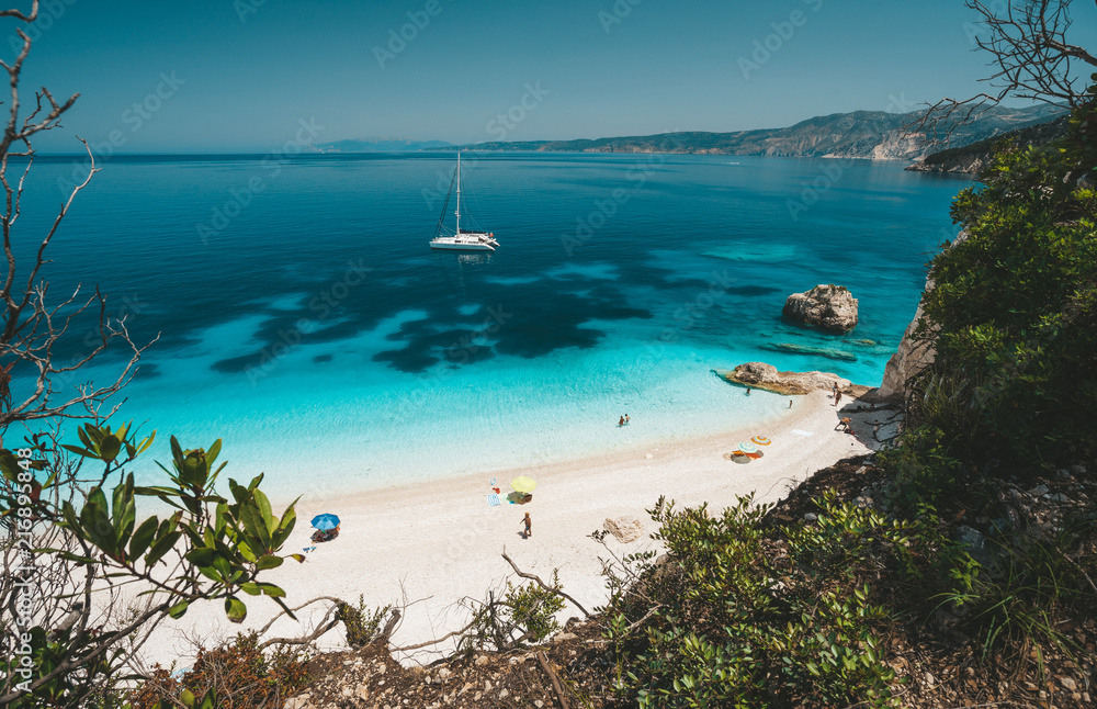 Fteri beach, Cephalonia Kefalonia, Greece. White catamaran yacht in clear blue sea water. Tourists on sandy beach near azure lagoon