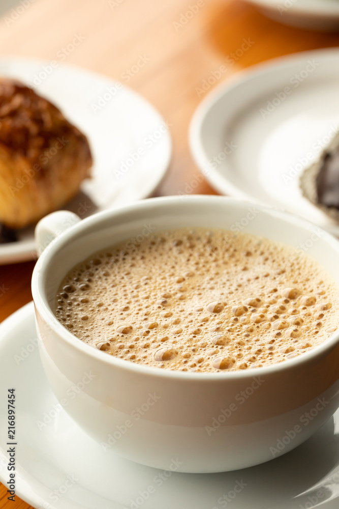 Close up on coffee in a white cup and saucer, with pastry in the background Close up on coffee in a white cup and saucer, with pastry in the background