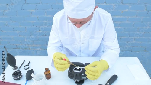 Elderly solid person in white dressing gown, cap, medical mask, glasses cleans matrix of digital camera with special means. Lens, sensor, matrix, cleaning wands, magnifier, isopropyl alcohol. photo