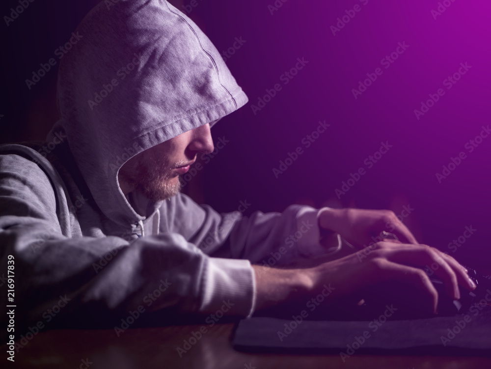 portrait of hooded man typing text on keyboard in front of pc monitor late in the night