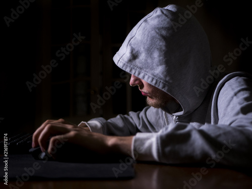 portrait of hooded man typing text on keyboard in front of pc monitor late in the night