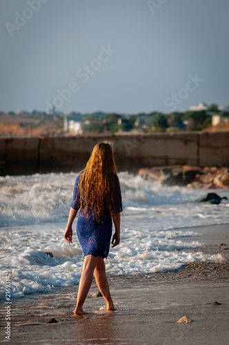 Beautiful natural blonde girl with long hair in short blue dress walking at sunset on the sea beach