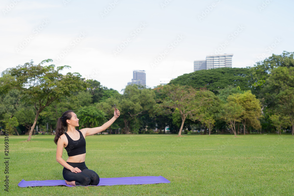 Woman Taking Selfie With Mobile Phone While Sitting On Yoga Mat In Park