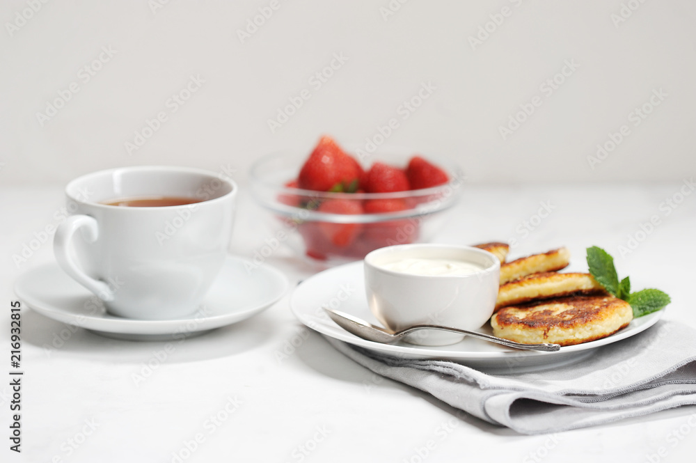 Table setting for breakfast. On a plate syrniki with sour cream. Next a cup of tea and a bowl of fresh strawberries. Light background. Close-up. Table setting for breakfast. On a plate syrniki with sour cream. Next a cup of tea and a bowl of fresh strawberries. Light background. Close-up.