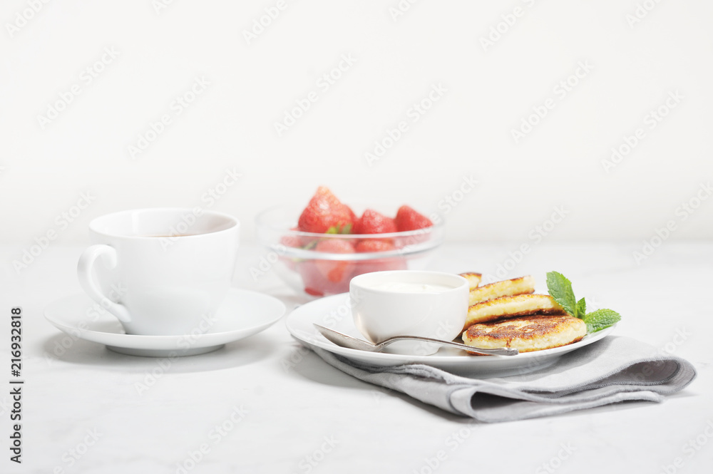Table setting for breakfast. On a plate syrniki with sour cream. Next a cup of tea and a bowl of fresh strawberries. Light background. Close-up. Table setting for breakfast. On a plate syrniki with sour cream. Next a cup of tea and a bowl of fresh strawberries. Light background. Close-up.
