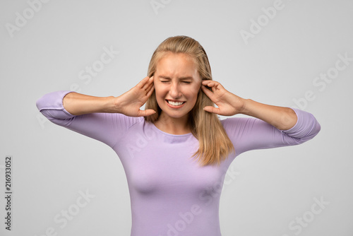 A young blonde woman frowns heavily while closing her eyes and clamping her head between her hands. Concept of loud unpleasant sound, headache, migraine, stress
